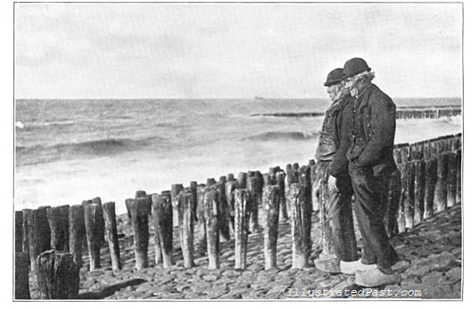 Two men look out to sea on the west coast of Holland. They are wearing Dutch clogs. The wooden stakes are to prevent erosion.