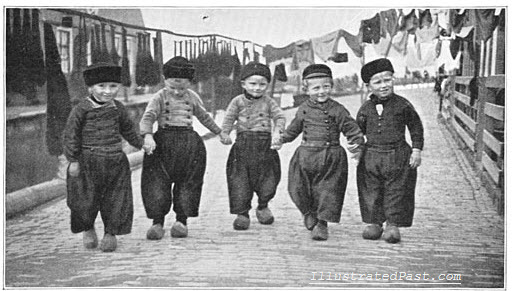 Dutch boys wearing clogs march down a quiet cobblestone street. Their clogs must have made quite the racket.