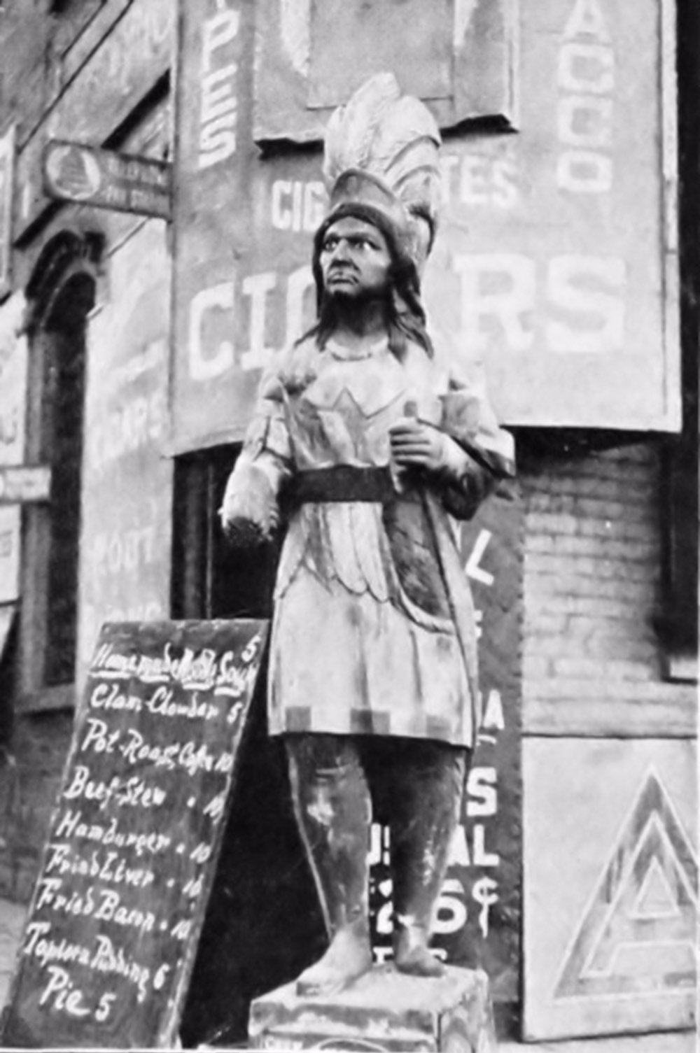 A Fearsome Cigar Store Warrior - Standing Next to a Blackboard Advertising a Restaurant Menu. The Statue is Missing a Hand.